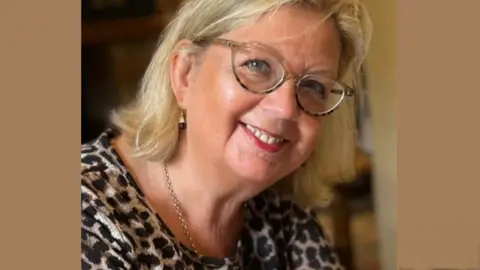 Family photo Close up shot of Janet Farmer smiling. She has shoulder length blonde hair and is wearing glasses and a patterned top.