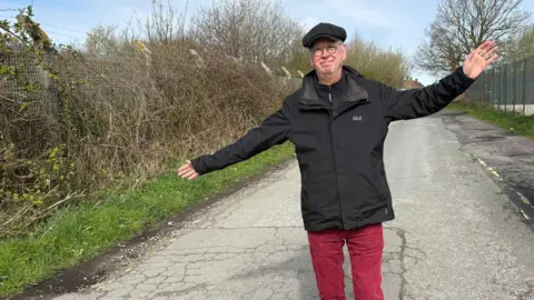 A middle-aged man wearing red trousers, a black jacket and a cap stands on an empty lane smiling with his arms outstretched. 