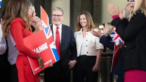 PA Media Keir Starmer and his wife Victoria smile as they walk through clapping crowds.