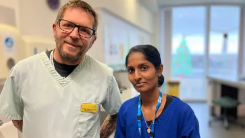 Nurse Sharifa wearing blue overalls and a lanyard with dark tied back hair, smiling at the camera, standing next to her colleague on the left