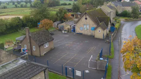 Aerial image of Hackforth and Hornby Church of England Primary School, showing two stone buildings and a playground, next to a single track road, with fields and trees in the background