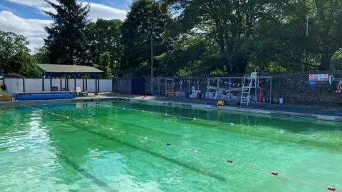 The outdoor pool has buoys attached to ropes to outline the lanes. There is colourful bunting on the stone wall to the right and bunting on a blue and grey veranda on the left. Work equipment is piled up behind a grey metal fence on in the corner.
