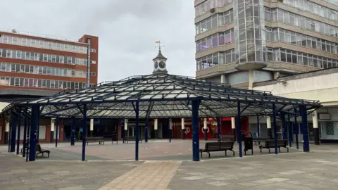 Anglia Square, a view of concrete paving, a central glass covered seating area and 1960s-style multi-storey office buildings in the background.