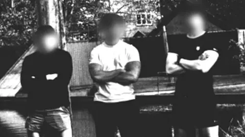 Members of Active Club North West pose in sportswear with their arms crossed in a rural setting for a group black and white photograph