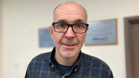 A close-up image of Angus Wilson. He is an older man with short, grey hair on the sides of his head and is bald on top. He is smiling at the camera. Angus is wearing a pair of black, square-framed glasses, and a blue and green plaid shirt.