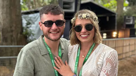 George King/BBC Craig Brown and Tara Sweeting are wearing sunglasses and green Latitude lanyards. They are standing close to each other and are smiling while looking into the camera.