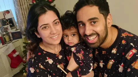 A couple hold their son between them as they smile at the camera. They all wear festive pyjamas and are in a room with a Christmas stocking on the wall. The woman has long dark hair past her shoulders, the man has short black hair and a short black beard. Their son looks to be young, toddler age.