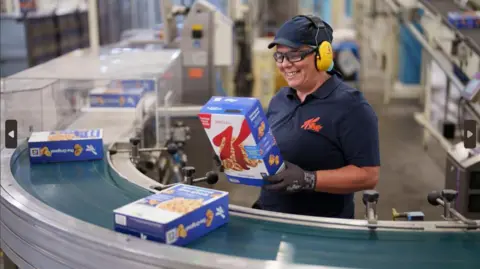 Kellogg's A woman in a navy Kellogg's top with a navy hat and yellow ear protectors. She is stood at the conveyor belt and she is picking up a Kellogg's box which is blue and there are two other boxes are on the belt.