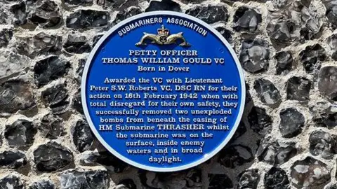 A blue commemorative plaque mounted on a flint stone wall details the bravery of Petty Officer Thomas William Gould VC.