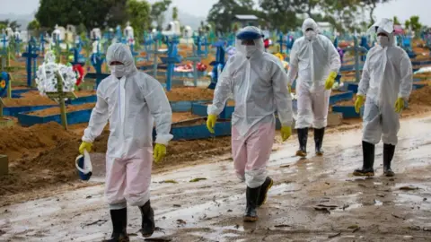 Getty Images Workers wearing protective suits walk past the graves of COVID-19 victims at the Nossa Senhora Aparecida cemetery, in Manaus, Brazil, on 25 February 2021.