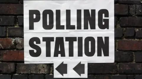 A white polling station sign with black letters and arrows pointing to the left. The sign is on a brick wall