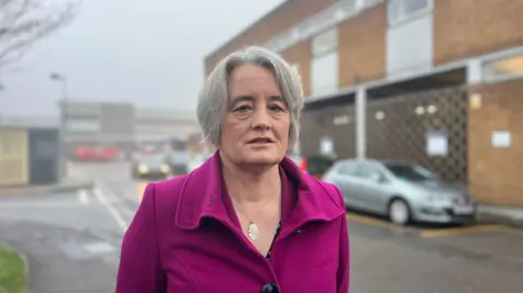 Claire Young MP standing outside the Royal Mail building in Yate, wearing a pink coat