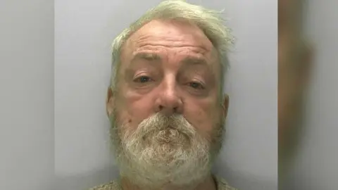 Mugshot of Edward Hanbury with white hair and a beard looking straight ahead. He is wearing a cream jumper and there is a grey background