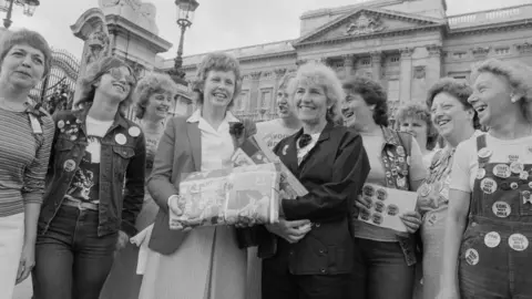 Getty Images Group of women lined up smiling and laughing in a black and white photo with the front of Buckingham Palace just visible behind them.