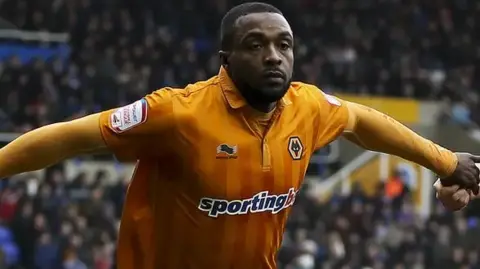 The footballer is on the pitch in a Wolves kit and has both arms outstretched. He has short hair and a beard. 