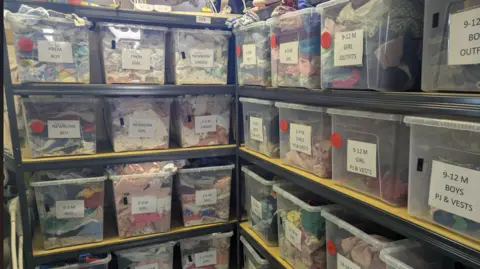 Emilie de Bruijn A small office space is full from ceiling to floor with boxes of different children's clothes. Signs on the front of the boxes identify who they are for 