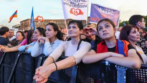 People, mostly women, stand behind a metal barrier looking ahead. One is a making a heart shape with her hands, and others are holding banners in Yerevan, Armenia on June 5. 