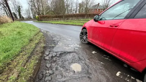 BBC A red car is driving on a rural road. There is a grass verge on one side and a red bricked house in the distance on the right hands side. There is a pothole filled with water on the left hands side beside the wheel of the car.