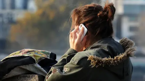 A generic stock photo of a woman using a mobile phone. The phone is a white colour and she is wearing a green coat with a fur hood.