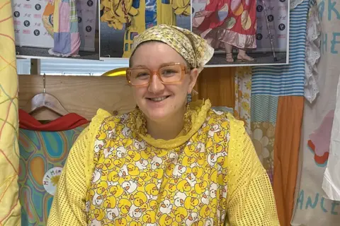 May Gauntlett in her workshop. She is wearing a variety of yellow, patterned clothing, including a headscarf. Fabric and design images are laid on the table and hanging behind her