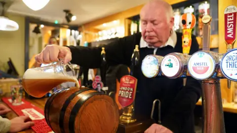 PA Media A man pouring a jug of bitter into a small barrel on a bar