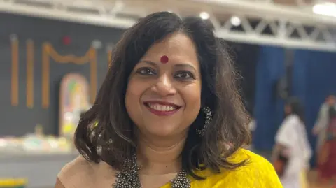 Barnali Ghosh is smiling at the camera, wearing traditional Indian dress in mostly yellow. In the background is a small stage with a shrine of a goddess on it.