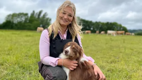 Harriet Cowan of the television series Clarkson's Farm sits in a field with a brown and white collie. She is wearing a dark coloured gilet and pink shirt. Her long blonde hair is blowing in the window. In the background, brown cows are grazing