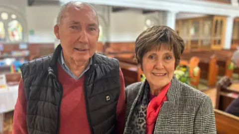 Bernard McPhee and his wife Doris smiling. They are standing in front of church pews in St George's. Doris is wearing black and white checked coat and pink scarf, and has short brown hair. Bernard is wearing a a red jumper, blue shirt and black gillet.