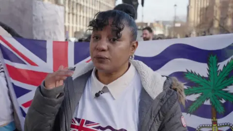 A woman stands in front a British Overseas Territory flag in Parliament Square. she has dark hair and is wearing a light coloured winter coat. 