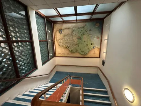 A large map on a wall above a staircase. It shows the old borders of County Durham with landmarks such as High Force waterfall and Durham Cathedral depicted.