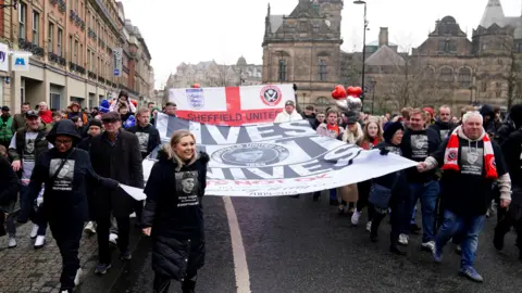 PA Media People marching holding a banner with Harvey Willgoose's face on it. There is another banner behind with Sheffield United and England badges on it. People are wearing black t shirts with Harvey's face on it.