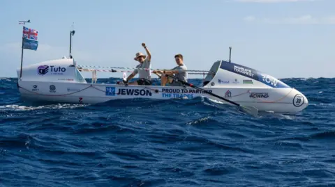 Two men on a white rowing boat on the ocean, with sponsors Jewson, Tuto and charity logo for Mind written on the side. The one at the front has his left hand raised and is grinning to the camera. He is wearing a hat and grey t-shirt. The man behind him is also looking to the camera, holding oars. He has dark brown hair.