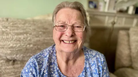 NIKKI FOX/BBC Dawn North, who was discharged back home in March after having a chest infection that followed a fall, looks at the camera, smiling. She has metal framed glasses and a light blue top with white flowers on it. She has greying hair and is sitting on a beige sofa in her living room. You can see her head and shoulders and there is a fireplace visible in the background.