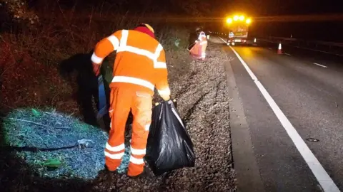 Two workers by the side of a main road wearing orange fluorescent outfits, collecting litter.