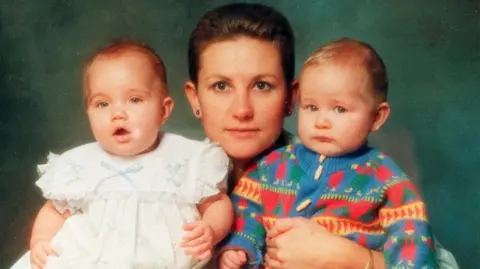 PA Media A photo from 1991 shows a woman with short hair and earrings with her arms around twins. The baby on the left is wearing a white dress, with the baby on the right dressed in a knitted baby grow. 