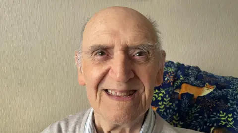 An image of Bob Dixon, he is an elderly bald man who is smiling.  He is wearing a shirt and grey jumper and is sitting in a chair with a blanket over his right should which has foxes and greenery on it.  It is a navy blanket.