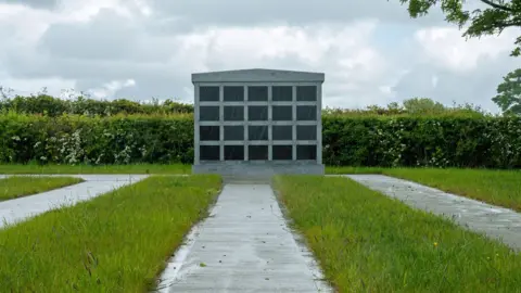 DCSDC A grey columbarium with a number of rows in a housed space.
It is near a grassy area with footpaths and shrubbery. 