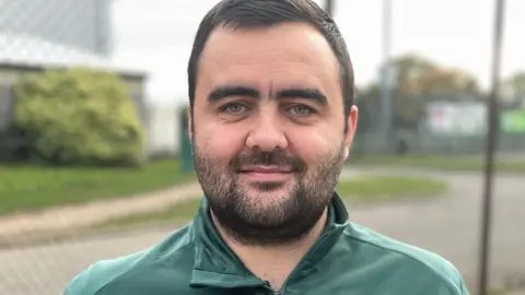 Mr Leeder wearing his Greenbank FC uniform in front of the closed pitches. 
He has a short beard and dark hair and his tracksuit is green with an emblem on his left chest.