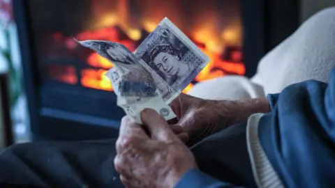 An older man's hands holding two twenty pound notes with an electric fire in the background.
