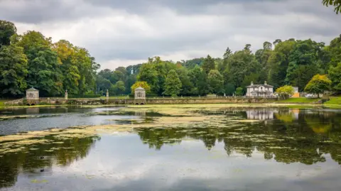 Getty Images A lake with a house and two smaller buildings in the background. Behind the buildings are trees.