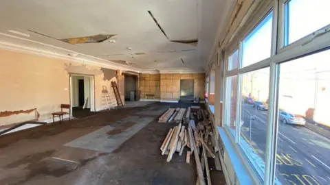 Auction Estates Interior of a room which has been stripped out. There is exposed plasterwork on the walls two visible walls, a damaged ceiling, and stacks of wood on the floor. to the right is a large window overlooking a road and some parked vehicles