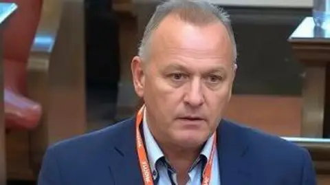 Darren Bamford Darren is wearing a blue suit jacket and shirt, without a tie, and an orange lanyard. He is picture in the council chamber as he is addressing councillors on his plea to keep Packmoor out of the local plan. 