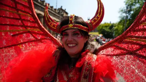 A parade participant wearing an elaborate red costume with horned headpiece and lace-like red wings poses close to the camera during a street festival.