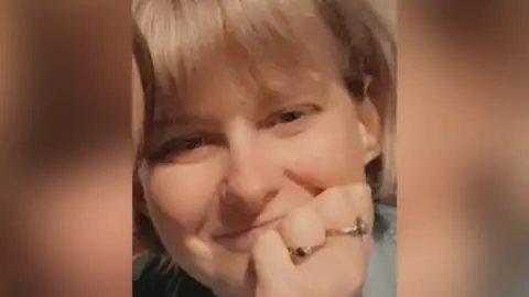 An image of Millie Mulcahy, she is a blonde white woman, smiling, she has her hand in front of her face and is wearing two rings.