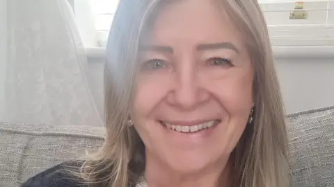 Anne Eason, one of the residents who had to leave the flats, sits on a sofa with a window behind her. She is wearing a cream top and black cardigan and has shoulder-length hair. She is smiling at the camera.