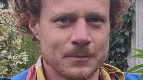 Ben Corser is smiling as he looks at the camera. He has blue eyes and light brown curly hair and a faint goatee. He is standing outdoors surrounded by plants. He is wearing a neck scarf.