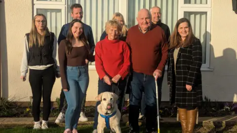 RSPCA The Potter family standing in the garden outside their home, with Daisy in front of them. There are two girls who look teenage, Rita and Philip centre, and two men and women in the family photo. The sun is shining.