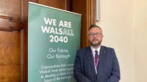 Pictured is Garry Perry with a slight smile on his face. He is wearing round dark framed glasses, has a dark blond quiff and a dark blonde beard. He is wearing a dark blue blazer with a white shirt underneath and a blue and red tie. He is standing next to a green sign that says "We are Walsall 2040".