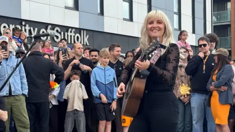 Zoie O'Brien/BBC Lianne Kaye smiling while performing with her guitar at Ipswich Waterfront. She has blonde hair and is looking towards the camera while strumming her guitar. She has crowds of people behind her.