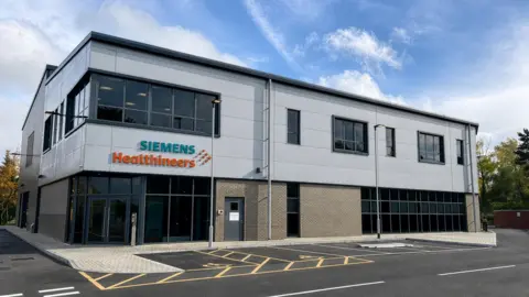 The modern two-storey commercial building with Siemens Healthineers branding displayed prominently on the front façade. The building is finished in white cladding and light brickwork.